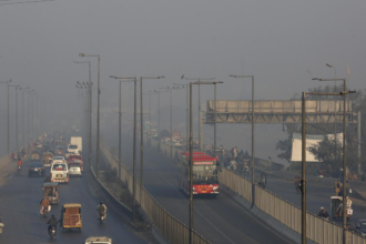 A smog-covered cityscape of Lahore with vehicles emitting smoke.
